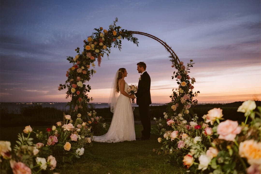 Bride and groom standing under a floral wedding arch at twilight with sunset sky