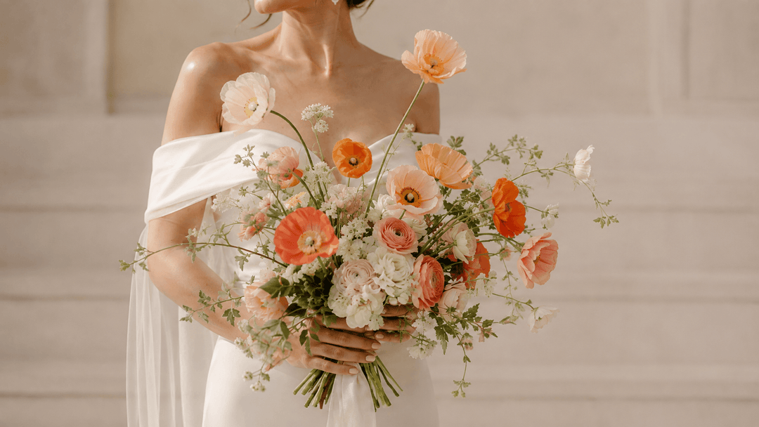 Bride holding a loose bouquet of peach, pink, white, and orange poppy wedding flowers with roses, ranunculus, and greenery.