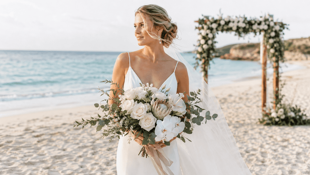 Bride holding resilient white beach wedding flowers beside an oceanfront ceremony arch.