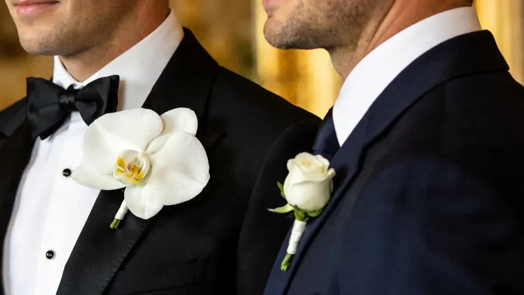 Editorial close-up of two men in tailored suits the groom in a black tux with a larger white real-touch silk orchid boutonniere, and the groomsman in a navy suit with a smaller white rosebud boutonniere.