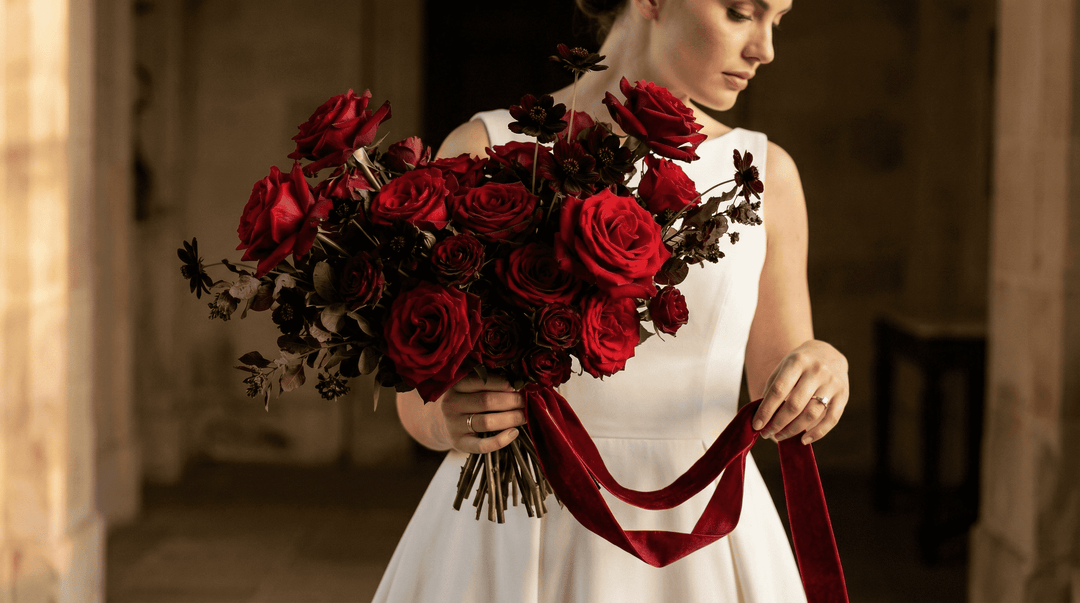 High-fashion, cinematic wedding photography. A close-up of a luxurious, deeply saturated red rose bouquet held against a heavy, matte crepe ivory wedding gown