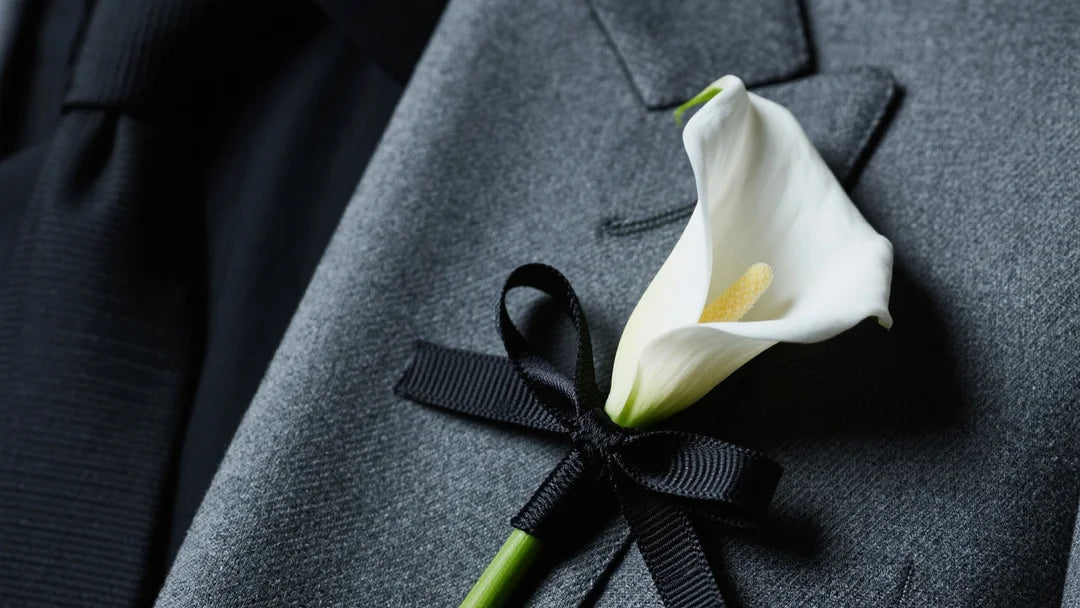 High-fashion close-up of a charcoal suit lapel with a minimalist real-touch white calla lily boutonniere, no greenery, stem wrapped in black silk ribbon.
