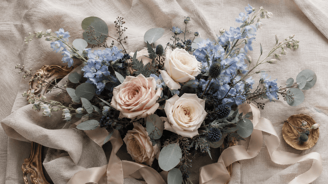 Luxury floral flat lay with dusty blue and blush flowers, ivory roses, delphinium, thistle, silver eucalyptus, and champagne ribbon.