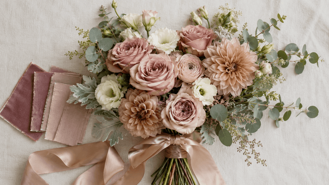 Luxury wedding flat lay featuring a dusty rose bridal bouquet on ivory linen with Quicksand roses, mauve garden roses, cream lisianthus, pale ranunculus, dahlias, eucalyptus, dusty miller, and champagne ribbon.