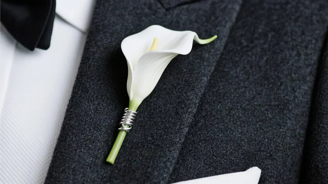 a sleek, pure white artificial miniature calla lily boutonniere pinned securely to a sharp, dark charcoal grey worsted wool tuxedo lapel